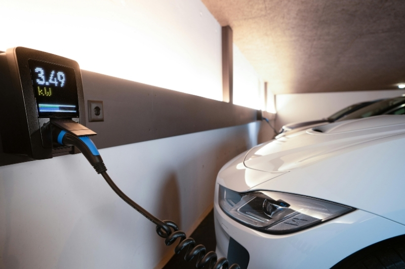 An electric car plugged into a wall-mounted home charging unit inside a garage, with the charging display visible.